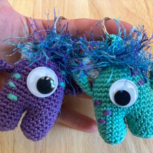 May include: Two handmade crochet monster keychains. One is purple with teal accents, the other is teal with purple accents. Both have large googly eyes and blue fuzzy hair. The keychains are small and whimsical.
