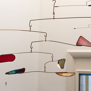 Puede incluir: Una escultura móvil con piezas de vidrio de colores que cuelgan de un marco de metal. Las piezas tienen varias formas, incluyendo un círculo, un triángulo y un rectángulo. La móvil está suspendida del techo.