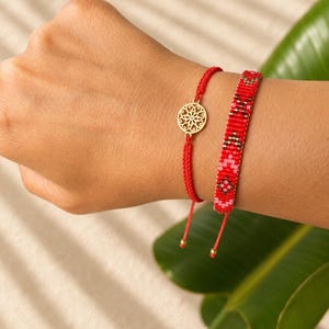 May include: A set of two red bracelets. One bracelet features a thin red cord with a circular gold-toned filigree charm. The second bracelet is a wider woven band made of red, pink, and metallic bronze beads arranged in a geometric pattern.
