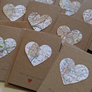 May include: A collection of brown paper envelopes with heart-shaped map cutouts. Each envelope has a different map location, including Leighton Buzzard, Denton, Wigan, and Ambleside. The envelopes have the text "Where our love began" printed on them.