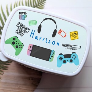 May include: White lunchbox with a blue, green and pink gaming theme. It features a Nintendo Switch, two game controllers, headphones, and the text "Level Up", "Game On", and "Harrison".