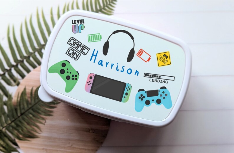 May include: White lunchbox with a blue, green and pink gaming theme. It features a Nintendo Switch, two game controllers, headphones, and the text "Level Up", "Game On", and "Harrison".