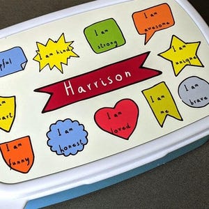 May include: A white and blue lunchbox lid decorated with colourful labels saying "I am helpful", "I am kind", "I am strong", "I am awesome", "I am unique", "I am brave", "I am smart", "I am funny", "I am honest", "I am loved", and "I am me". The name "Harrison" is on a red banner. A green apple is beside it.