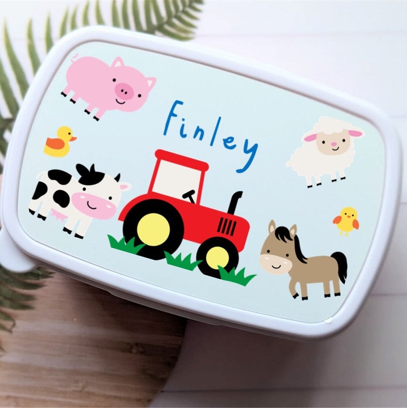 May include: A white lunchbox with a blue background and a farm theme. The lunchbox features a red tractor, a cow, a pig, a sheep, a horse, and a chick. The lunchbox is personalised with the name "Finley".