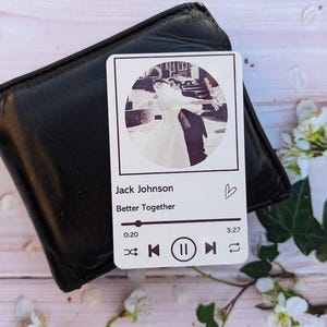May include: A black leather wallet with a white card featuring a black and white photo of a couple dancing. The card has the text "Jack Johnson Better Together" and a music player interface design. The background includes white flowers.
