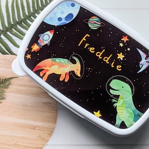 May include: A white lunchbox with a black background featuring a space-themed design. The lunchbox has a colourful dinosaur, a rocket ship, stars, planets, and the name "Freddie" written in yellow.