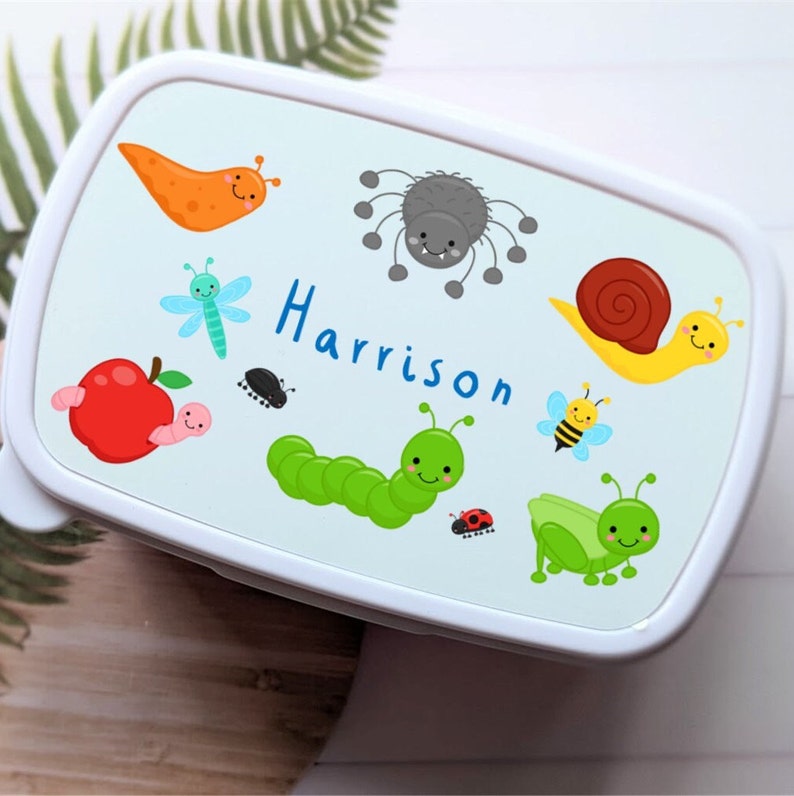 May include: White lunchbox with a blue background and colourful bug illustrations. The lunchbox has the name "Harrison" written on it.
