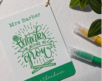 Pode incluir: Um cartão verde e branco com o texto "Mrs. Barber thanks for helping me grow love Theodore" e um desenho verde de uma flor e lápis.
