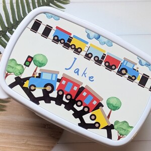 May include: White lunchbox featuring a colourful train design. The train is on a track with a red traffic light and green trees. The lunchbox is personalised with the name "Jake".