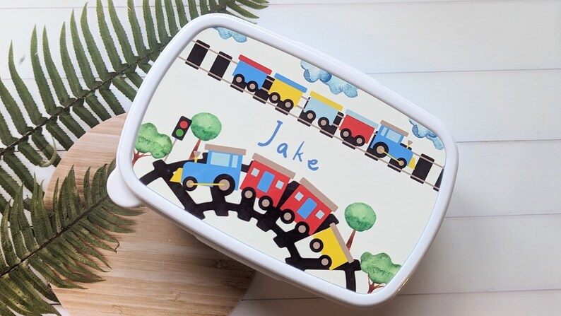 May include: White lunchbox featuring a colourful train design. The train is on a track with a red traffic light and green trees. The lunchbox is personalised with the name "Jake".