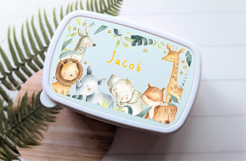 May include: A blue lunchbox with a white border and a jungle animal theme. The lunchbox features a lion, zebra, rhino, elephant, tiger, and giraffe. The name "Jacob" is written in yellow letters on the front of the lunchbox.