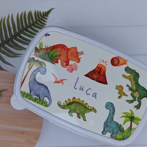 May include: White lunchbox featuring a colourful dinosaur design. The lunchbox has a blue, green, orange and red dinosaur design with a volcano and a comet. The lunchbox is personalised with the name "Luca".