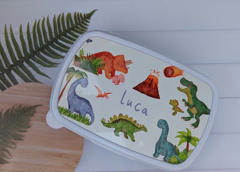 May include: White lunchbox featuring a colourful dinosaur design. The lunchbox has a blue, green, orange and red dinosaur design with a volcano and a comet. The lunchbox is personalised with the name "Luca".