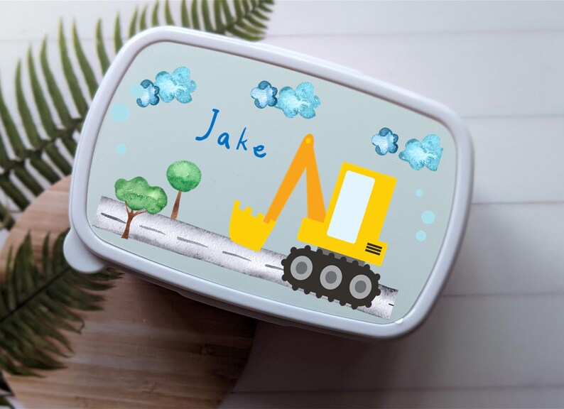 May include: A blue lunchbox featuring a yellow excavator design. The excavator has a yellow cab and black wheels. The lunchbox is personalised with the name "Jake".
