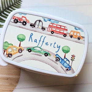 May include: White lunchbox featuring a colourful cartoon design of cars, trucks, and a double-decker bus on a winding road. The name "Rafferty" is written in blue cursive lettering.