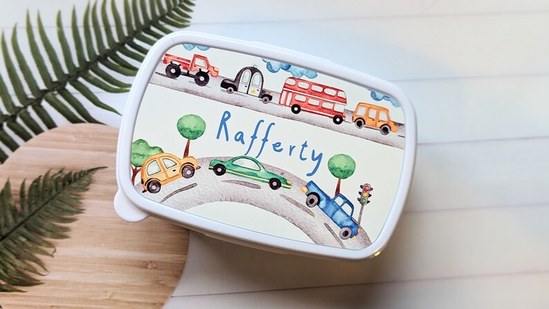 May include: White lunchbox featuring a colourful cartoon design of cars, trucks, and a double-decker bus on a winding road. The name "Rafferty" is written in blue cursive lettering.
