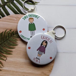 May include: Two white round keychains with a cartoon illustration of a boy and a girl. The boy is wearing a green shirt and the girl is wearing a purple shirt. The keychain for the boy says "Jake Bilenciz Langley Park" and the keychain for the girl says "Gracie H Year 4".