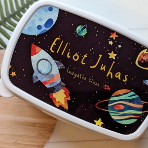 May include: A white lunchbox with a black background featuring a rocket, planets, and stars. The lunchbox is personalised with the name "Elliot Juhas" and the text "ladybird class".