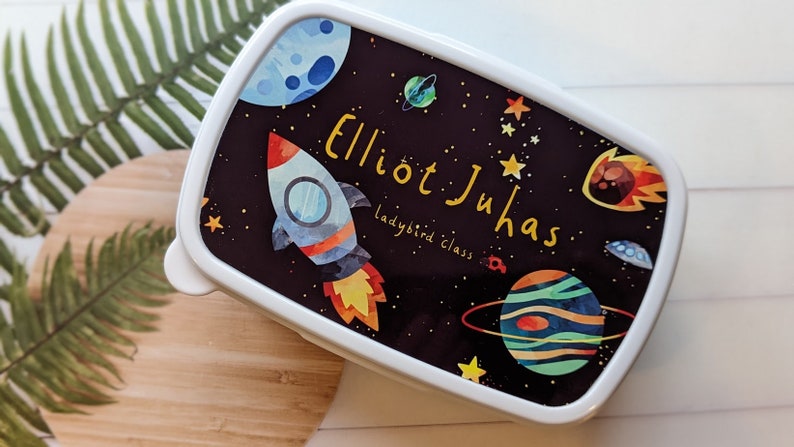 May include: A white lunchbox with a black background featuring a rocket, planets, and stars. The lunchbox is personalised with the name "Elliot Juhas" and the text "ladybird class".
