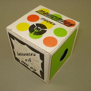 Puede incluir: Una caja blanca con un patrón de lunares verdes, amarillos y naranjas. La caja tiene un logotipo en blanco y negro con un tenedor y el texto "MAGIC BOX" y "ПРОФЫ". La caja también tiene una etiqueta negra con el texto "Играшески куб МК-02".
