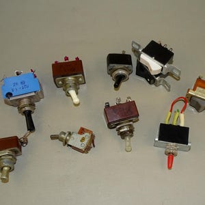Può includere: Una collezione di vari interruttori a levetta, con diversi colori e design. Gli interruttori hanno corpi neri, marroni e blu, con leve rosse, bianche e nere. Alcuni hanno cablaggio e hardware di montaggio visibili. Per applicazioni elettriche.