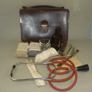 Op de afbeelding: Een vintage medische kit met een bruine leren koffer, een stethoscoop, verbanden en andere medische benodigdheden. De stethoscoop heeft een rode slang en een zwart oordopje. De kit bevat een oogbad en een bruine fles.