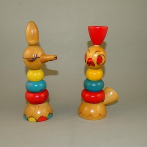 Puede incluir: Dos juguetes de apilar de madera con forma de pájaros. El pájaro de la izquierda tiene un pico largo y está pintado de amarillo, azul, rojo y verde. El pájaro de la derecha tiene un sombrero rojo y está pintado de amarillo, azul, rojo y verde.