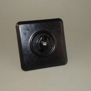 Könnte beinhalten: Ein schwarzer quadratischer Lichtschalter mit einem runden, schwarzen Knopf in der Mitte. Der Knopf hat ein stilisiertes Flammenmotiv.