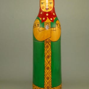 May include: A traditional Russian nesting doll, or matryoshka, painted in a folk art style. The doll is made of wood and features a woman in a red headscarf and green dress with yellow geometric patterns.