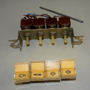 May include: An assortment of vintage automotive switches and indicator lights. The switches are mounted on a metal bracket with toggle levers. The indicator lights are rectangular with various symbols, including headlights and a circle with a line.