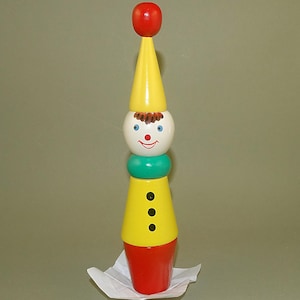 Puede incluir: Un juguete de payaso de madera pintado con colores brillantes. Presenta una parte superior roja, un sombrero y cuerpo cónicos amarillos, una cara blanca sonriente, un cuello verde y tres botones negros.