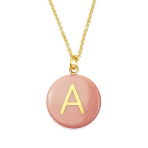 Può includere: Una collana con catena dorata con un ciondolo rotondo rosa con la lettera A in oro.