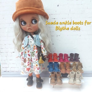 Puede incluir: Una muñeca con un vestido floral, chaleco vaquero y sombrero marrón, con una colección de botines de ante de varios colores. Las botas se exhiben en una pequeña estantería blanca. El texto de la imagen dice: "Suede ankle boots for Blythe dolls."
