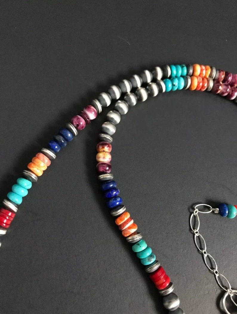 May include: A multi-colored beaded necklace with a silver chain clasp. The beads are made of turquoise, lapis lazuli, coral, and other stones. The necklace is a beautiful example of Native American jewelry.