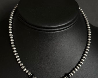 Könnte beinhalten: Eine silberne Perlenkette mit schwarzen Perlen, die durchgehend dazwischen liegen. Die Kette befindet sich auf einer schwarzen Puppe.