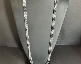 May include: A long silver necklace with multiple strands of small beads. The necklace is displayed on a black mannequin and is laying in a loop on the table.