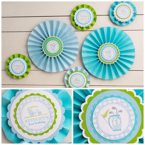 May include: A selection of blue and green paper fan decorations with golf-themed centrepieces. The centrepieces feature golf cart and golf club illustrations, with text that reads "A Hole in One" and "Anthony's first birthday."