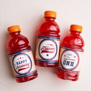 May include: Three red juice bottles with baseball-themed labels. The labels are brown with blue and red accents. One label says "Happy Birthday", one says "Mason", and one says "One".