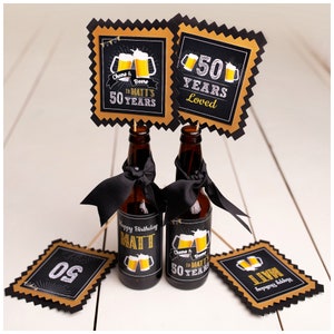 May include: Two small beer bottles with black ribbon and labels that say "Happy Birthday Matt" and "Cheers & Beers to Matt's 50 Years".  There are four black and gold signs with the numbers "50" and the text "50 Years Loved".