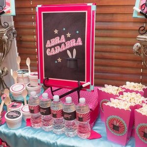May include: A magic-themed party table setting with pink and blue decorations. There are various treats, including popcorn, ice cream toppings, and water bottles. A chalkboard sign reads "Abra Cadabra" with a magician's hat and rabbit ears.