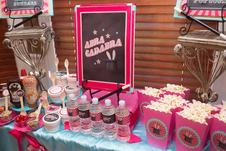 May include: A magic-themed party table setting with pink and blue decorations. There are various treats, including popcorn, ice cream toppings, and water bottles. A chalkboard sign reads "Abra Cadabra" with a magician's hat and rabbit ears.