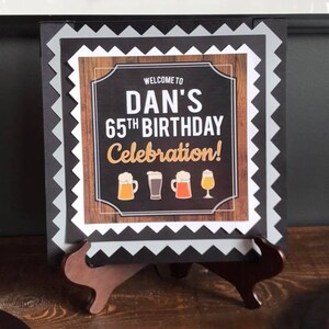 May include: A black and white sign with a woodgrain background and a white border. The sign says "Welcome to Dan's 65th Birthday Celebration!" with a row of beer mugs below the text.