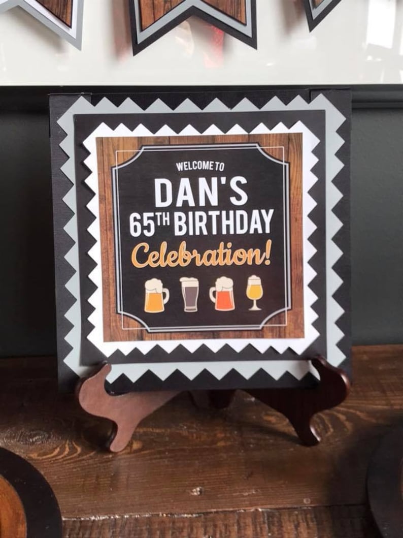 May include: A black and white sign with a woodgrain background and a white border. The sign says "Welcome to Dan's 65th Birthday Celebration!" with a row of beer mugs below the text.