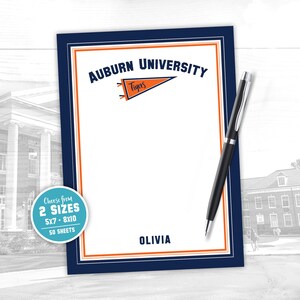 May include: A personalised notepad with the text "Auburn University" and a pennant reading "Tigers". The notepad has a navy blue border with orange accents and includes the name "Olivia". A black pen rests on the notepad.
