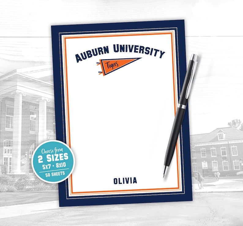 May include: A personalised notepad with the text "Auburn University" and a pennant reading "Tigers". The notepad has a navy blue border with orange accents and includes the name "Olivia". A black pen rests on the notepad.