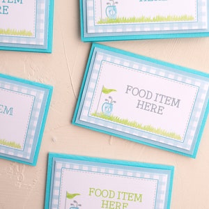 May include: Four blue and white gingham food labels with a golf bag and golf clubs graphic. The text on the labels reads "Food Item Here".