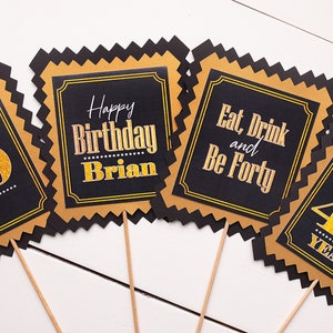 May include: Four black and gold square signs with gold glitter text on black backgrounds. The signs say "40", "Happy Birthday Brian", "Eat, Drink and Be Forty", and "Cheers to 40 Years".