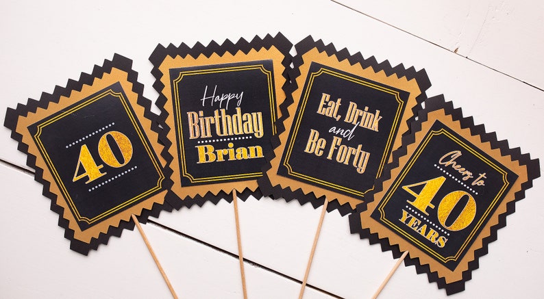 May include: Four black and gold square signs with gold glitter text on black backgrounds. The signs say "40", "Happy Birthday Brian", "Eat, Drink and Be Forty", and "Cheers to 40 Years".