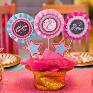 May include: A pink and blue table centerpiece with three paper rosettes, each with a different name, "Anna Gabriela", "Rose", and a number "8". The rosettes are attached to sticks and placed in a gold glitter top hat with pink tissue paper. The centerpiece is surrounded by gold glitter and pink and blue stars.