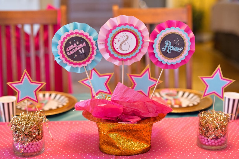 May include: A pink and blue table centerpiece with three paper rosettes, each with a different name, "Anna Gabriela", "Rose", and a number "8". The rosettes are attached to sticks and placed in a gold glitter top hat with pink tissue paper. The centerpiece is surrounded by gold glitter and pink and blue stars.